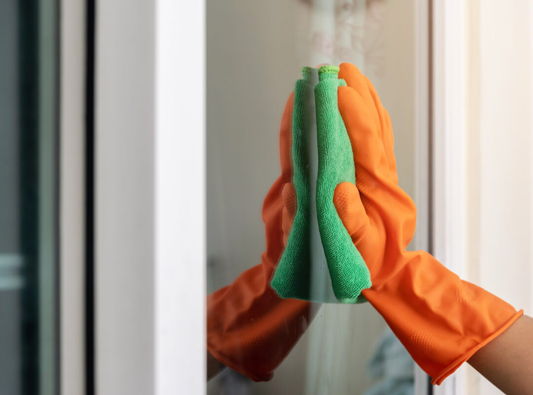 Women's hand wears orange rubber gloves, using green cloth to wipe the glass of the building with a glass cleaner.