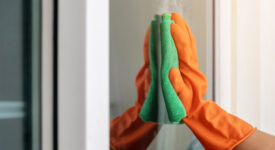 Women's hand wears orange rubber gloves, using green cloth to wipe the glass of the building with a glass cleaner.