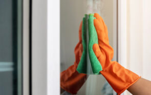 Women's hand wears orange rubber gloves, using green cloth to wipe the glass of the building with a glass cleaner.