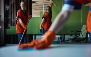 Smiling cute cleaner with a mop in her hands posing for the camera in the presence of her busy colleagues