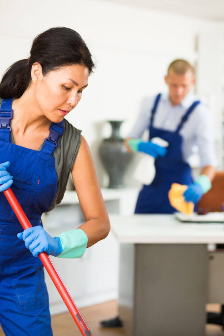 Portrait of professional female worker of cleaning service wearing uniform and rubber gloves wiping office floor with mop