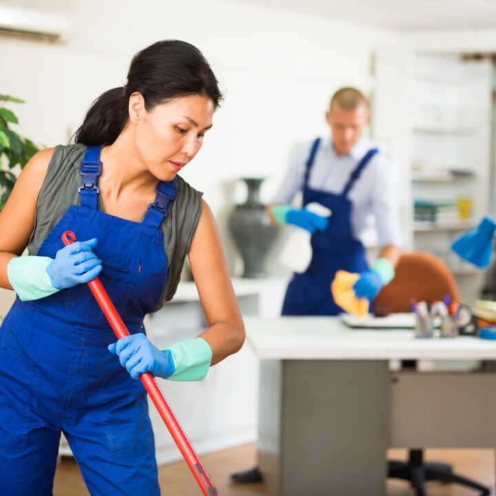 Portrait of professional female worker of cleaning service wearing uniform and rubber gloves wiping office floor with mop