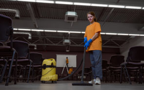 Full-size photo of focused young janitor vacuum-cleaning the floor in the conference hall