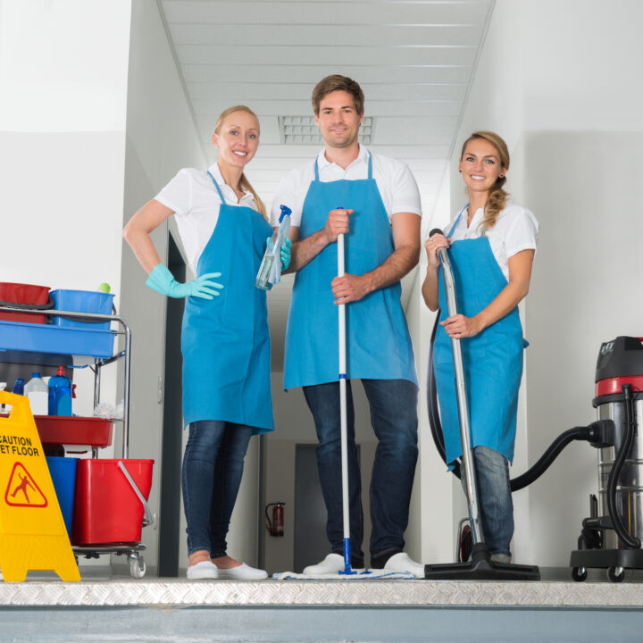 Portrait Of Smiling Janitors Holding Cleaning Equipments By Yellow Wet Caution Sign On Floor